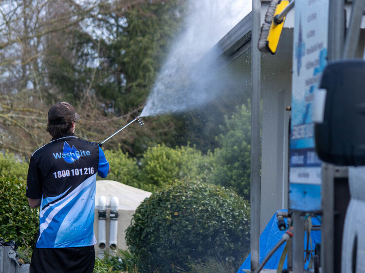 Worker pressure washing the exterior of a house surrounded by greenery.